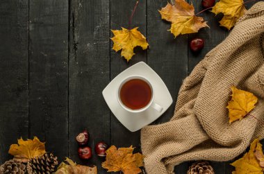 Sıcak çay, sıcak örgü kış battaniyesi, sonbahar yaprakları, kestane ve koni. Düz görünüm kompozisyonu siyah ahşap arkaplan üzerine yatıyordu.