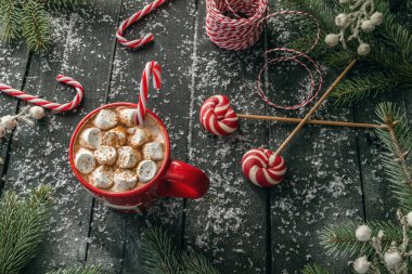 Bir kupa sıcak Noel kakaosu, üzerine tarçın ve şeker kamışları serpiştirilmiş. Ağaç dalları, hediye paketleme ipleri ve lezzetli lolipoplarla çevrili..