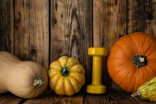 Orange autumn pumpkin with yellow dumbbell. Healthy fitness lifestyle rustic fall composition for Halloween or Thanksgiving. Gym workout and training concept, with gourds, squash types.