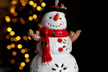 Seramik kardan adam heykelciği. Noel ve kış için güzel, şenlikli bir oyuncak süslemesi, arka planda ağaç ışıkları var..