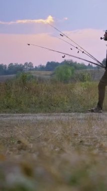 Sakallı orta yaşlı bir adam gün batımında açık bir alanda yürür omzunda makaralı birkaç olta ile. Koyu renk tişört, kamuflaj sırt çantası ve bej panama giyiyor.