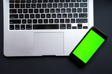 Laptoplu modern siyah ofis masası ve yeşil renkli ekranlı akıllı telefon. Ofis işi ve iş kavramı. Üst Manzara. Düzleştirme biçimi.