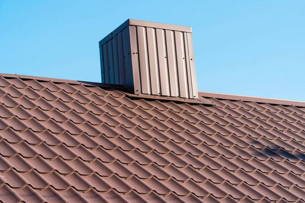 The roof of a house with modern metal tiles and a chimney, against the background of a blue sky. Metal roofing.