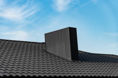 Modern, siyah bir baca, siyah, fayanslı bir çatının üzerinde oturur. Baca dikey çizgilerle dikdörtgendir. Arka plan beyaz bulutlu mavi bir gökyüzü..