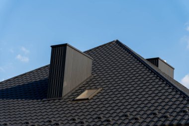 Çağdaş bir evin çatısında, mimari zarafet ve dayanıklılığı vurgulayan parlak mavi gökyüzüne karşı koyu renkli fayanslar ve iki belirgin baca bulunur..