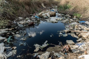 Plastik molozlarla dolu kirli bir dere gökyüzünü yansıtıyor, çöpün doğal su kütlesi üzerindeki etkisini gösteriyor, acil çevresel kaygıları vurguluyor.