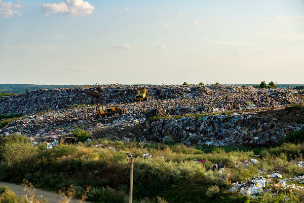 The landscape shows a sprawling landfill with heavy machinery working among heaps of refuse. Green vegetation partially surrounds the area, illuminated by afternoon sunlight.
