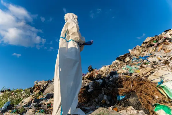 stock image A waste management worker in protective clothing inspects a landfill filled with various types of debris under a sunny blue sky in the daytime.