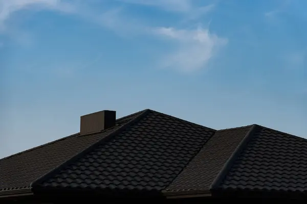 stock image The roof of a suburban house is visible against a bright blue sky with wispy clouds. The textured tiles create a distinct pattern, enhancing the architectural features.