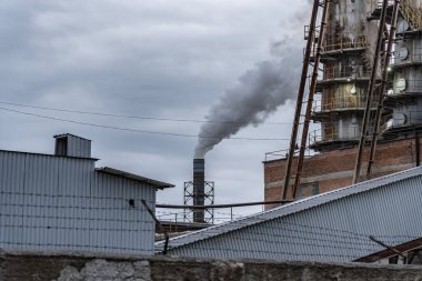 Endüstriyel yapılar ve çatılarla çevrili bulutlu gökyüzüne duman salan bir fabrika, kentsel bir çevreyi gözler önüne seriyor..