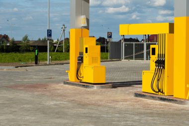 Parlak, modern bir benzin istasyonunda açık mavi gökyüzünün altında geniş ve açık bir alanda sarı pompalar bulunur. Yakın park yerleri görülebilir, erişilebilirliği arttırıyor.