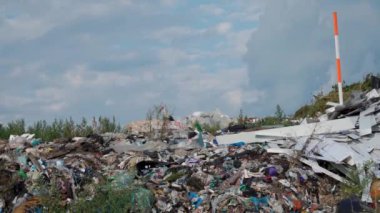 Bulutlu bir gökyüzünün altında çeşitli çöp türleriyle dolu geniş bir çöp sahası gösteriliyor. Etrafa enkaz saçılmış durumda, yakınlarda da biraz yeşillik var. Yüksek kalite 4k görüntü