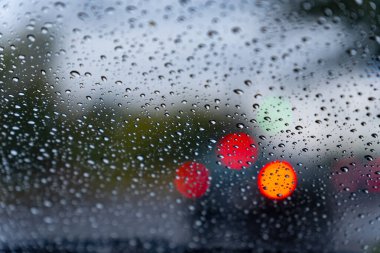 Yağmur damlaları pencereyi kaplar, arka plandaki araçların kırmızı ışıklarını bulanıklaştırır, tipik bir yağmurlu gün atmosferini betimler..