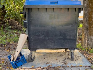 Mavi tepeli bir çöp bidonu sakin bir parkta duruyor, yanında atılmış bir karton ve plastik bir sandık var, doğadaki günlük atık sorunlarını vurguluyor..