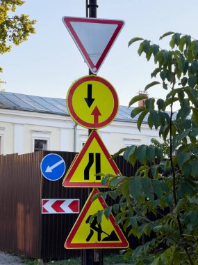 Renkli uyarı işaretleri, açık mavi gökyüzünün altındaki yoğun bir kavşakta hem yayaları hem de araçları yönlendiren çeşitli trafik yönetmeliklerini gösteriyor..