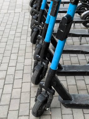 Canlı mavi scooter 'lar hareketli bir şehir kaldırımında hazır bekliyor, enerjik sürücüler güneşli bir günde şehir manzarasını keşfetmeye davet ediyorlar..