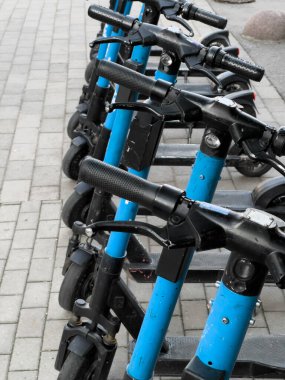 Bir sıra mavi scooter tuğla kaldırıma park edilmiş. Mobiletlerin hepsi aynı yöne bakıyor ve hepsi yan yana park edilmiş. Sahne düzen ve düzen hissi veriyor..