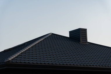 Modern bir evin çatısında parlak siyah metal fayanslar, berrak mavi gökyüzü altında çağdaş inşaat teknikleri ve malzemeler vurgulanıyor..