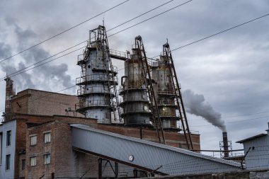 Yoğun, karanlık duman bulutlu gökyüzü ve motorlu makinelerle çevrili bir endüstriyel kompleksin yüksek bacalarından yükselir ve aktivite belirtileri gösterir..