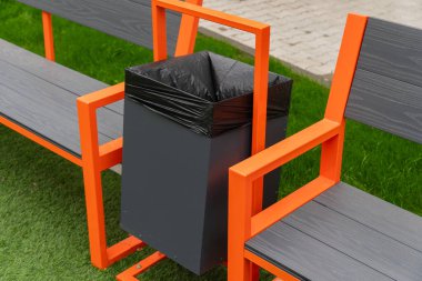 Siyah plastik kaplamalı siyah bir çöp tenekesi turuncu bir bankın üzerine monte edilir. Banka gri ahşap kapaklı metalden yapılmıştır. Bankta yeşil bir çimenlik var..