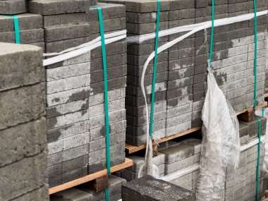 Gri kaldırım taşları ahşap paletlere düzgünce istiflenmiş, yeşil kayışlarla korunmuş. Bu tuğlalar şehir kaldırımında kurulmayı bekliyor, dış manzarayı geliştirmeye hazır..