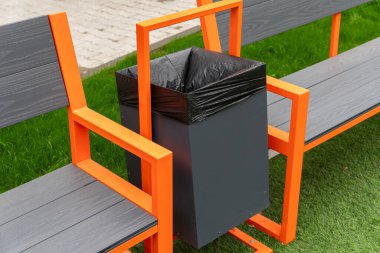 Canlı bir parkta, iki turuncu bankın arasına parlak bir çöp konteynırı monte edilir, ziyaretçileri temiz bir çevrede çöpleri güvenli bir şekilde atmaya teşvik eder..