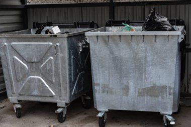 İki metal çöp konteyneri yan yana duruyor, biri siyah çöp torbası ve çeşitli atıklarla dolu, diğeri ise boş, alacakaranlıkta bir çöp toplama sahasında..