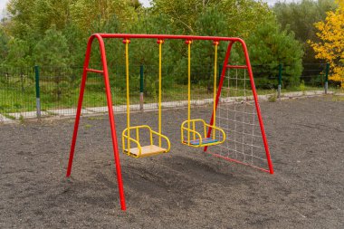 İki boş salıncak halka açık bir parktaki parlak kırmızı çerçevede asılı, çocukların sallanmaktan ve oynamaktan zevk almalarına hazır. Yeşillik daha canlı bir zemin hazırlıyor..