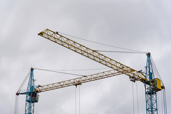 Two large construction cranes are actively positioned at a construction site, lifting materials as workers prepare the framework for a new house. The sky above is overcast.