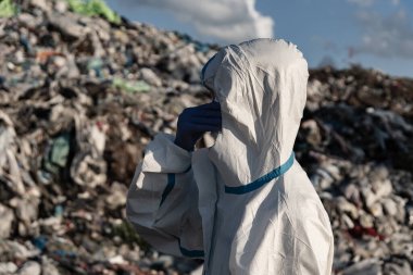 Koruyucu giysili bir işçi, parçalı bulutlu bir gökyüzünün altındaki atık yığınlarıyla çevrili geniş bir araziyi inceliyor. Çevre güvenliğinin önemi çok açık..