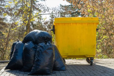 Parlak sarı bir çöp tenekesi, güneşli bir günde düşüşün renklerini gösteren ağaçların arka planına kurulmuş koyu plastik poşetlere karşı tezat oluşturuyor..