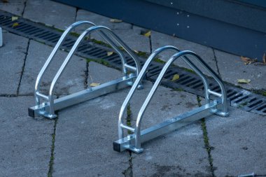 İki parlak metal bisiklet askısı kaldırımlı bir yüzey üzerinde duruyor, bisikletlere güvenli park alanı sağlamak için hazırlar. Gösterişli tasarım şehir çevresine modern bir dokunuş katıyor..