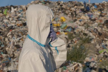 Koruyucu giysili bir işçi açık bir gökyüzünün altında çöp ve plastik atıkla dolu geniş bir alanı araştırıyor. Bu kirlilik ve ekoloji sorunlarını vurgular.