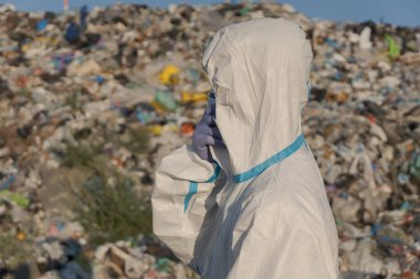Koruyucu ekipman kullanan bir atık yönetimi çalışanı, kirliliği önleme ve ekosistem sağlığına odaklanan plastik ve diğer atıklarla dolu büyük bir çöp sahasının yanında duruyor. Sağlık hizmetleri açıkça görülüyor..