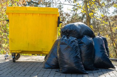 Toplumdaki atık yönetimi ve çevresel koruma çabalarının altını çizen siyah çöp torbaları, halka açık bir alanda canlı sarı bir çöp konteynırının yanına istiflenmiştir..