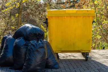 Siyah çöp torbaları ağaçlık bir alanda canlı sarı bir çöp konteynırının yanına istiflenir. Atık yönetimi ve çevre koruma alanının önemli bir yönünü gözler önüne serer..