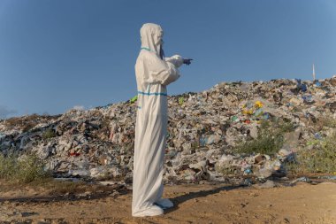 Koruyucu giysili bir insan çöplükte duruyor ve büyük bir çöp yığınını işaret ediyor. Çevre alanı, insan faaliyetlerinden kaynaklanan çevresel kirliliğin boyutunu ortaya koymaktadır..