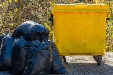 Siyah çöp torbaları bir parkta parlak sarı bir çöp kutusunun yanına istiflenir. Çevredeki ağaçlar, çevre temizliği ve uygun atık imhası ihtiyacını vurguluyor..