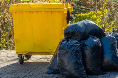 Siyah çöp torbaları açık hava ortamında sarı bir çöp konteynırının yanına dizilir. Sahne, kentsel bir ortamda atık tasfiye uygulamalarını vurguluyor.