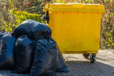 Dışarıdaki asfalt bir yüzeyde birkaç siyah çöp torbasının yanında sarı bir çöp konteyneri duruyor. Sahne, toplumdaki uygun atık imhası ve geri dönüşüm çabalarının önemini yakalıyor..