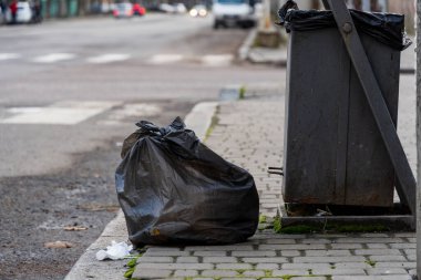 Siyah bir çöp torbası geri dönüşüm kutusunun yanındaki kaldırımda duruyor ve günlük kentsel atık yönetimi çabalarını gösteriyor. Sahne, yoldan geçen araçlarla tipik bir şehir caddesini yakalıyor.