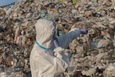 Koruyucu giysi giyen bir temizlik işçisi plastik atık ve molozlarla dolu geniş bir atık alanına el kol hareketi yapar..