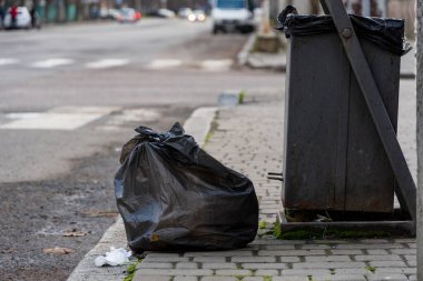 Şehir kaldırımı boyunca uzanan metal çöp konteynırının yanında duran iki siyah çöp torbası, kentsel atık imhası ve çevre koruma çabalarıyla devam eden sorunları vurguluyor.