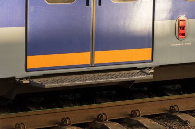 Bir tren istasyonuna yerleştirilmiş modern bir elektrikli tren kapısının yakın çekimi. Parlak renkler, gün batımında demiryolu rayları ve çevre ile tezat oluşturarak çarpıcı bir görsel yaratıyor..