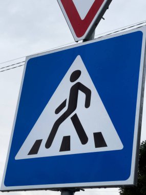 Üçgen mavi uyarı işareti, inşaat bariyerleri arasında yürüyen ve bir çalışma alanındaki yaya varlığına dair sürücülere bilgi veren bir kişiyi gösteriyor. Bu işaret işlek bir kavşağa yerleştirilmiştir.