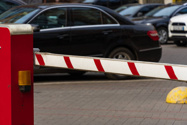 A bright red barrier stands at the entrance of a busy parking lot, preventing entry while vehicles wait in line. The scene reflects measures for safety and security in public spaces.
