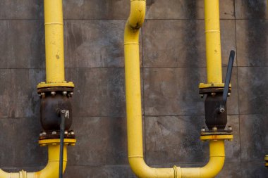 Sarı gaz boru hatları yıpranmış gri bir duvar boyunca uzanarak modern mühendislik ve endüstriyel estetiğin bir karışımını ortaya çıkarıyor. Vanalar, enerji sisteminin karmaşık dizaynını yansıtan bağlantılar yaratır..