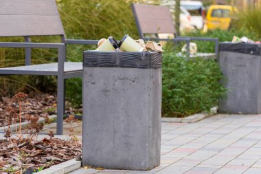 Parlak bir şehir parkında güneşli bir öğleden sonra, çöp torbalarıyla dolu beton bir çöp konteynırı.