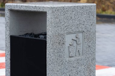 Modern bir gri çöp konteyneri şehir bölgesinde duruyor, yoldan geçenleri çöplerini güvenli bir şekilde atmaya davet ediyor. Tasarım basit işaretlerle geri dönüşüm ve çevresel farkındalığı teşvik ediyor..