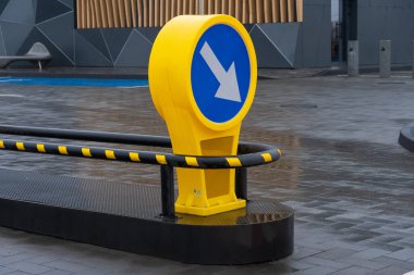 Çağdaş bir şehir kavşağında sol tarafında ok işaretli parlak sarı bir işaret. Islak asfalt çevreyi yansıtıyor, kentsel tasarımı ve yağmurlu havayı vurguluyor..